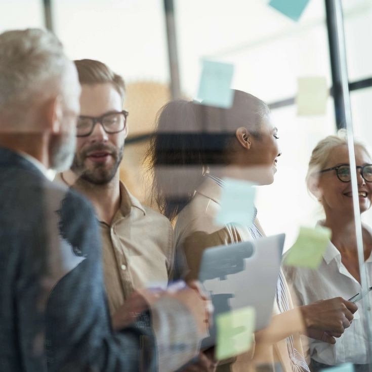 Mehrere Kolleginnen und Kollegen stehen vor Glaswänden mit Haftnotizen und besprechen Ideen.