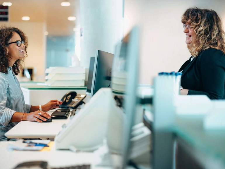 Mitarbeiterin am Serviceschalter spricht lächelnd mit einer Besucherin in einem modernen Büro.