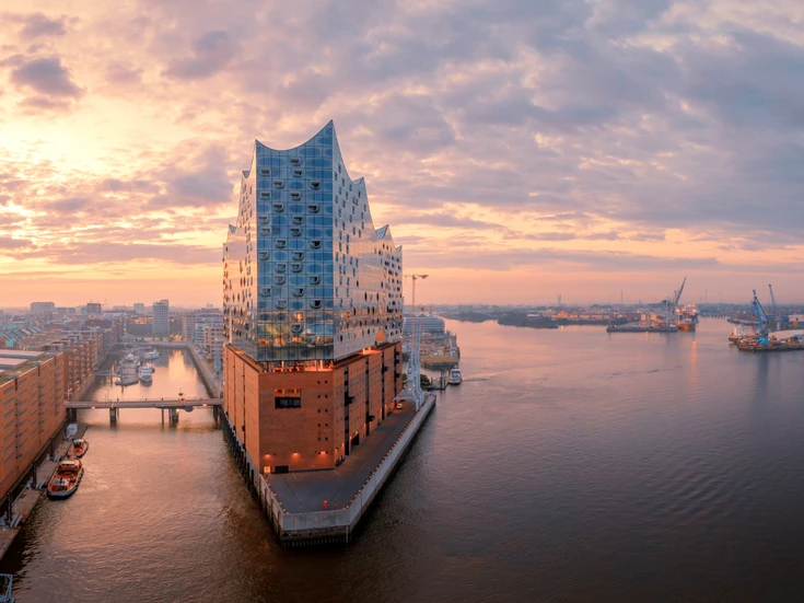 Elbphilharmonie in Hamburg at sunset, modern glass building on a brick base by the harbor, surrounded by water, bridges, and industrial structures under a colorful evening sky