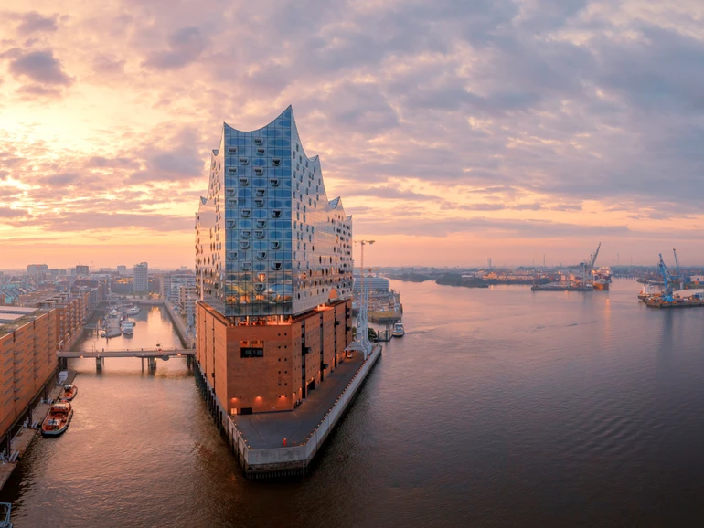 Elbphilharmonie in Hamburg bei Sonnenuntergang, modernes Glasgebäude auf Backsteinsockel am Hafen, umgeben von Wasser, Brücken und Industrieanlagen unter farbigem Abendhimmel