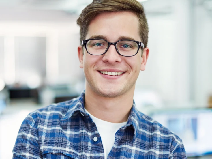 Portrait im Büro Junger Mann mit Brille im karierten Hemd lächelt in die Kamera, heller Büroraum im Hintergrund