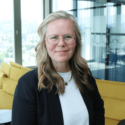Porträt einer lächelnden Frau mit Brille und langen, blond-braunen Haaren, die in einem modernen Büro vor einer gelben Couch steht.