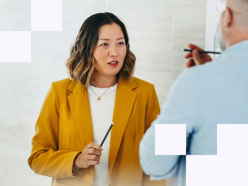 Frau in gelbem Blazer spricht mit einer Person und hält einen Stift in der Hand