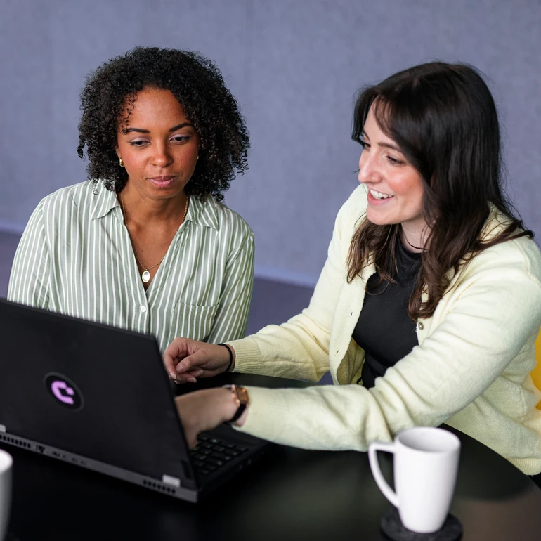 Zwei junge Frauen in gestreifter Bluse und gelbem Cardigan schauen auf den Bildschirm eines Laptops