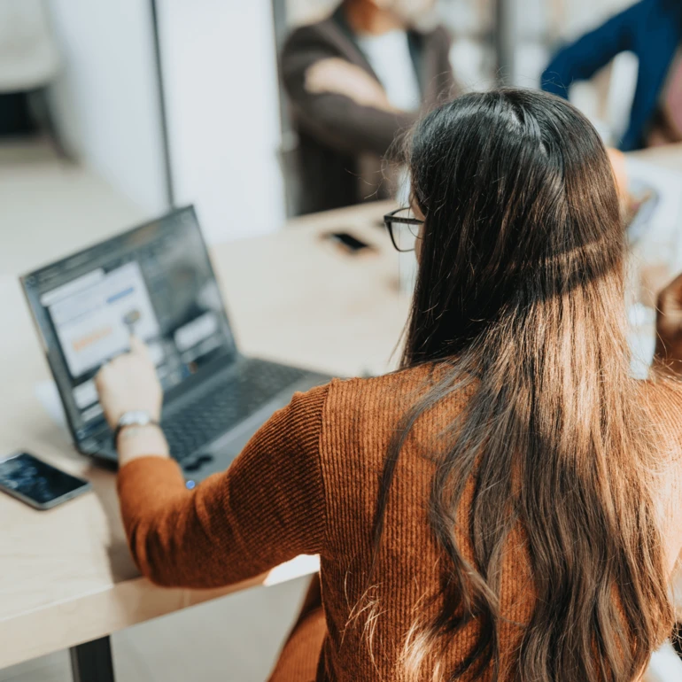 Junge Frau sitzt an einem Schreibtisch und arbeitet an einem Laptop, während andere Personen im Hintergrund unscharf zu sehen sind.