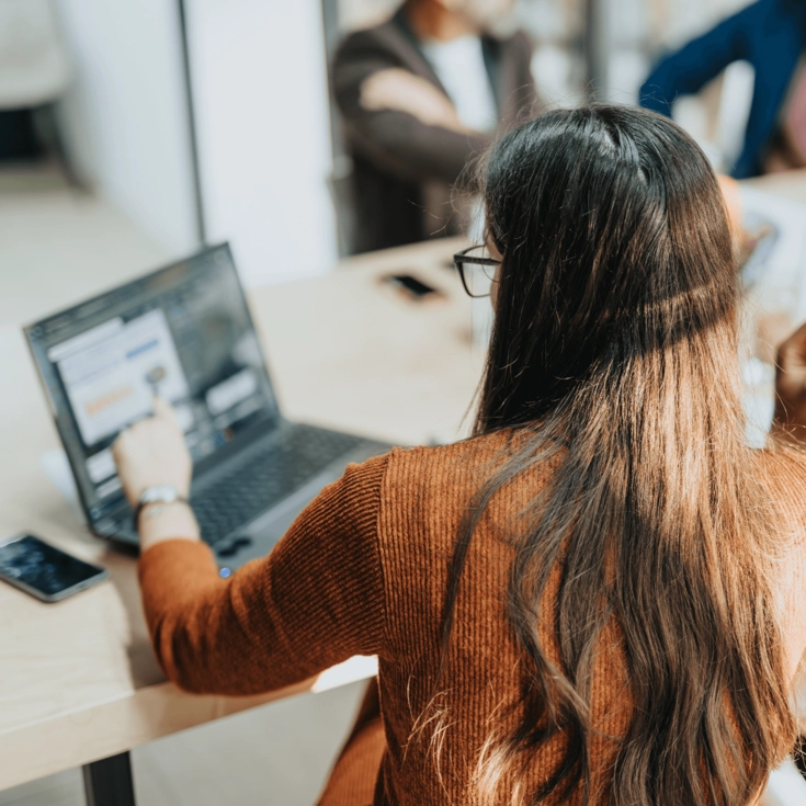 Junge Frau sitzt an einem Schreibtisch und arbeitet an einem Laptop, während andere Personen im Hintergrund unscharf zu sehen sind.