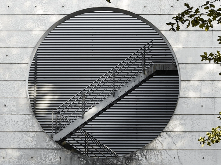 Modernes Gebäude mit kreisförmiger Öffnung und einer diagonalen Metalltreppe vor lamellenartiger Fassade.