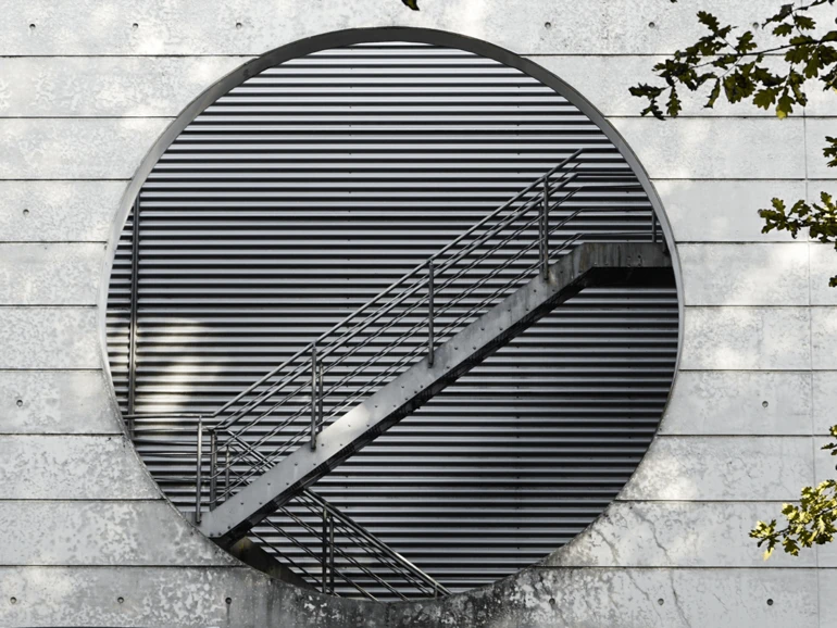 Modernes Gebäude mit kreisförmiger Öffnung und einer diagonalen Metalltreppe vor lamellenartiger Fassade.