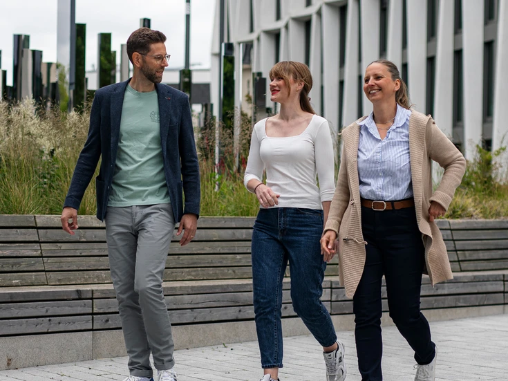 Drei Personen, ein Mann und zwei Frauen, laufen locker vor dem Bürogebäude und lächeln sich an