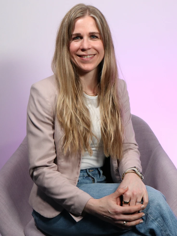 Portrait einer lächelnden conet Mitarbeiterin in beigem Blazer sitzend in Studio auf hellbraunem Stuhl
