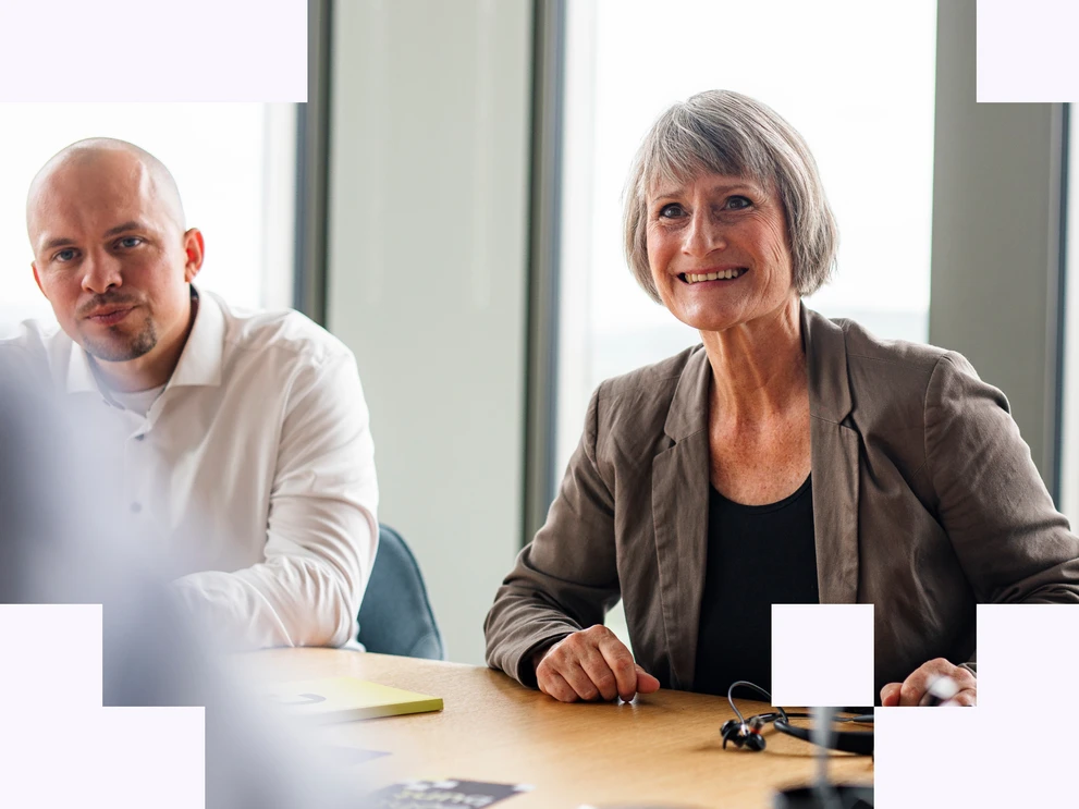 Frau mit dunklem Shirt und braunem Blazer in offenem Austausch im Meeting mit Mann in weißem Hemd