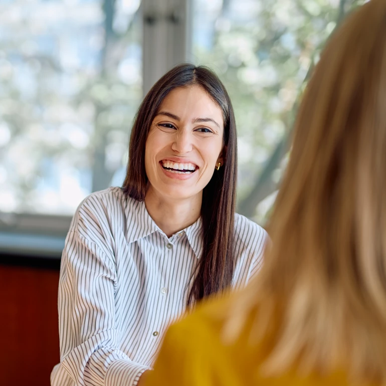 Gespräch im Büro Junge Frau mit langen dunklen Haaren im hellblauen Hemd lacht im Gespräch mit einer Person im Vordergrund