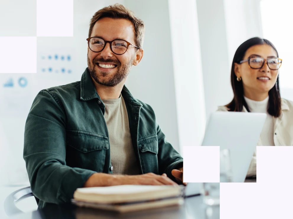 Mann mit offenem grünen Hemd, Brille und Bart sowie eine Frau mit weißem Blazer und Brille lachen gemeinsam vor einem Laptop in einem Meeting
