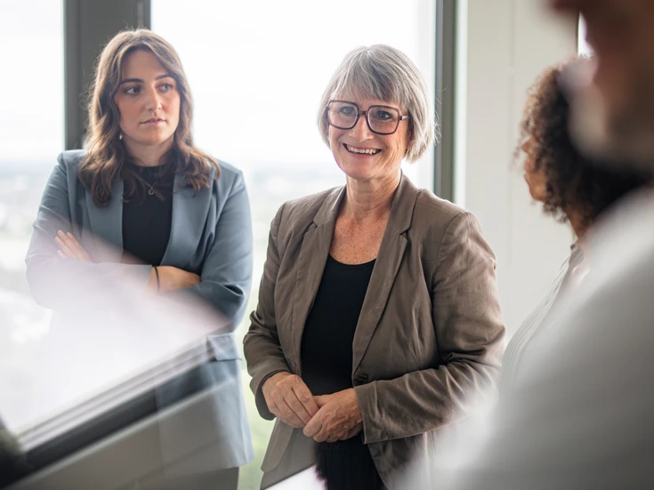 Frau in braunem Blazer und dunkler Brille steht in Meeting und lächelt