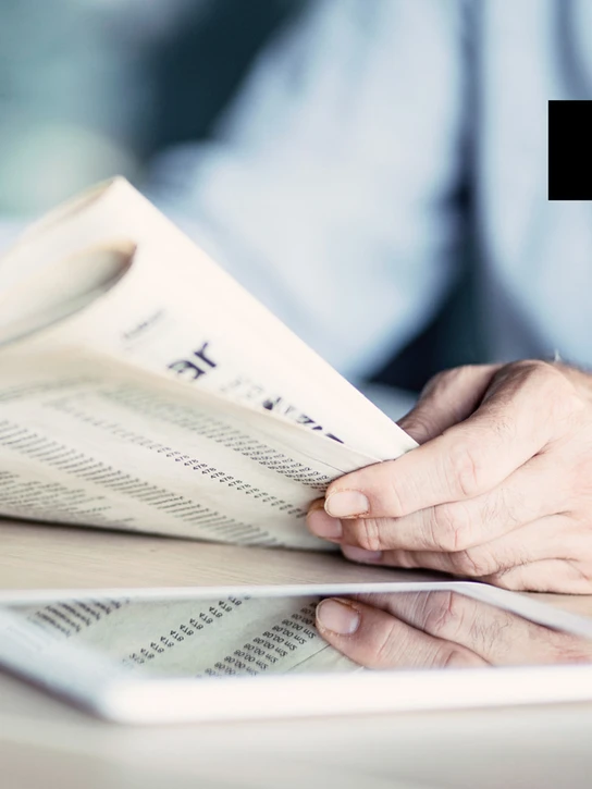 Foto: Nahaufnahme – Person blättert in Zeitung, die sich in Tablet spiegelt