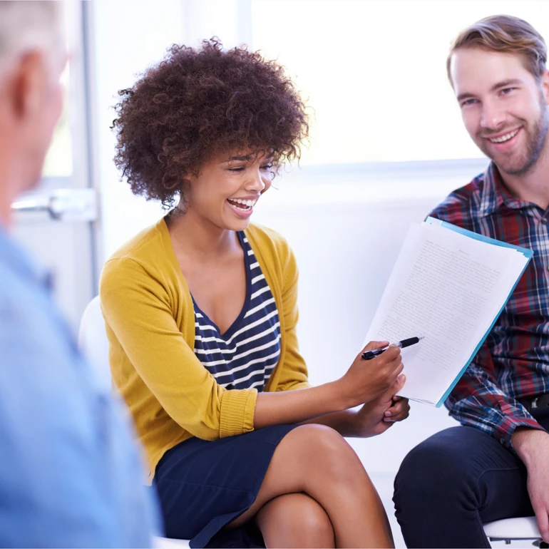 Frau in gelbem Cardigan und Mann im karierten Hemd sitzen im Gespräch, die Frau hält ein Dokument in der Hand