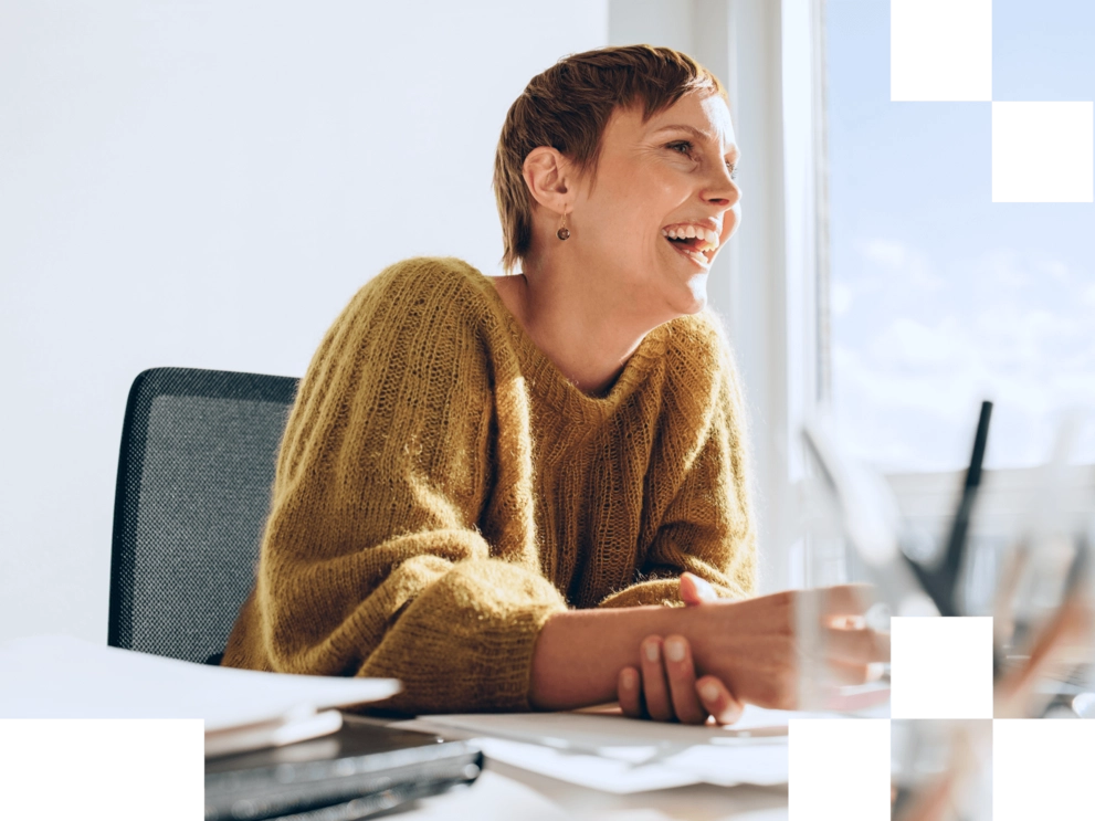 Lachende Frau sitzt in einem hellen Büro an einem Schreibtisch und wirkt entspannt und motiviert.