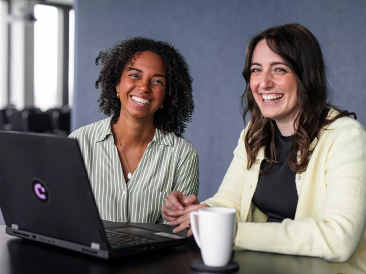 Teamwork im Büro Zwei junge Frauen in gestreifter Bluse und gelbem Cardigan sitzen vor einem Laptop und lachen