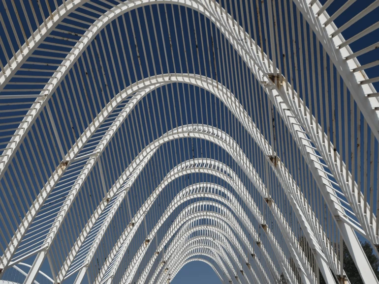 Blick auf die moderne weiße Bogenkonstruktion des Olympiageländes in Athen vor blauem Himmel.
