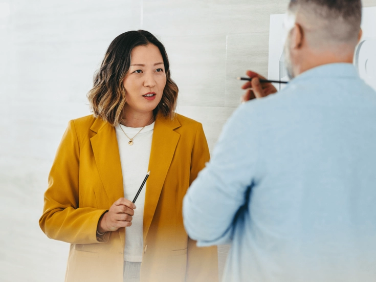 Frau im gelben Blazer spricht mit einem Kollegen und hält einen Stift, als würde sie eine Idee erklären.