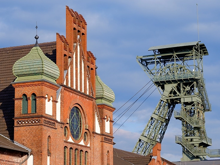 Brick facade of a historic building at the Zollern coal mine in Dortmund, featuring green ornamented domed towers and a large arched circular window; next to it rises a steel winding tower against a blue sky.