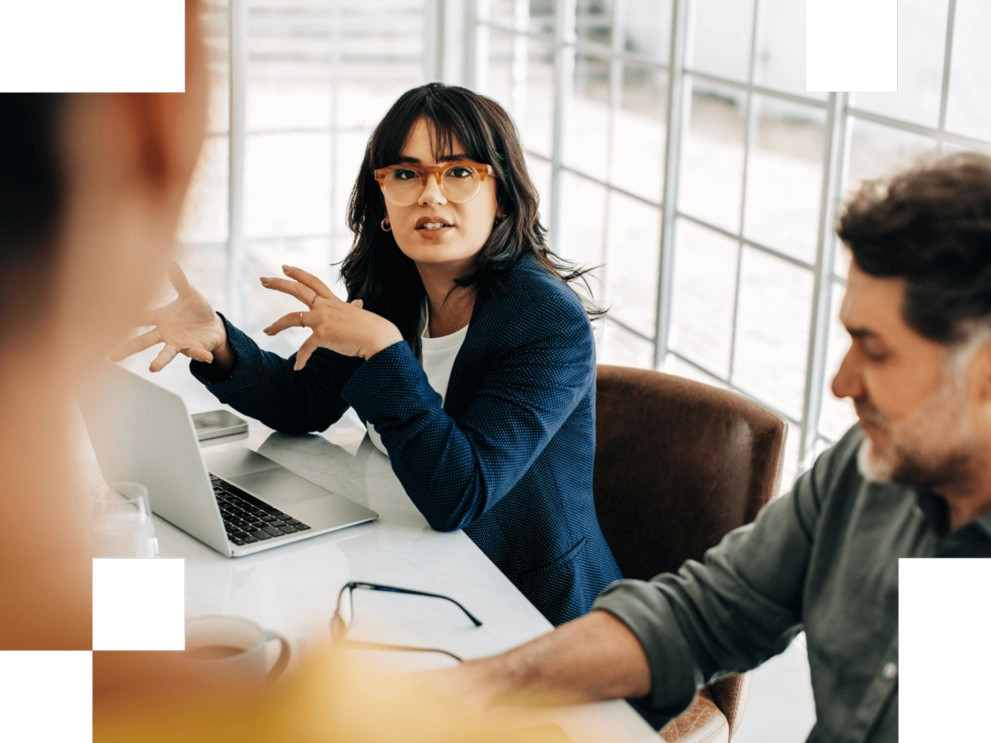 Eine Frau mit Brille und dunkelblauem Blazer spricht engagiert in einem Meeting, vor ihr ein Laptop; daneben sitzt ein Kollege.