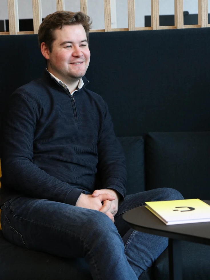 Portrait eines lächelnden conet Mitarbeiters in schwarzem Pullover locker sitzend auf Sofa mit gelbem Kissen