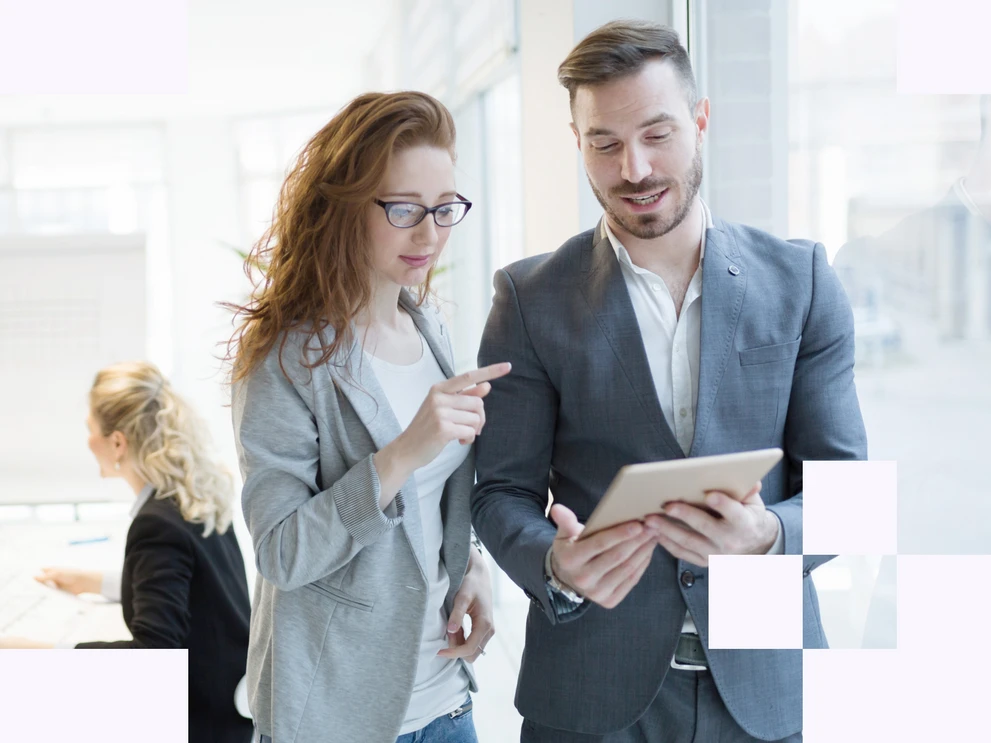 Foto mit zwei Personen, die im Büro zusammen stehen und ein Tablet betrachten während im Hintergrund weitere Personen an einem Arbeitsplatz arbeiten.