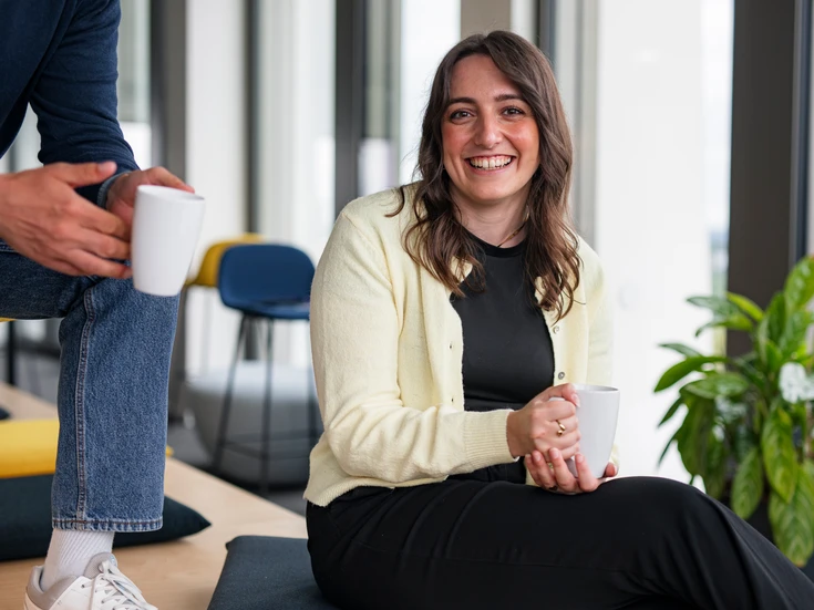 Junge Frau in gelbem Cardigan sitzt lachend im Open Space im Büro