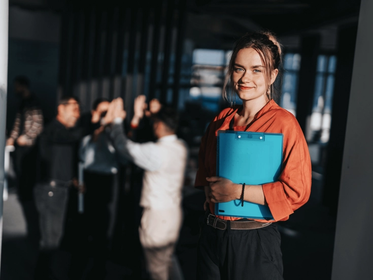 Junge Frau mit blauem Klemmbrett steht lächelnd im Vordergrund, während im Hintergrund eine Gruppe von Menschen zusammenarbeitet.