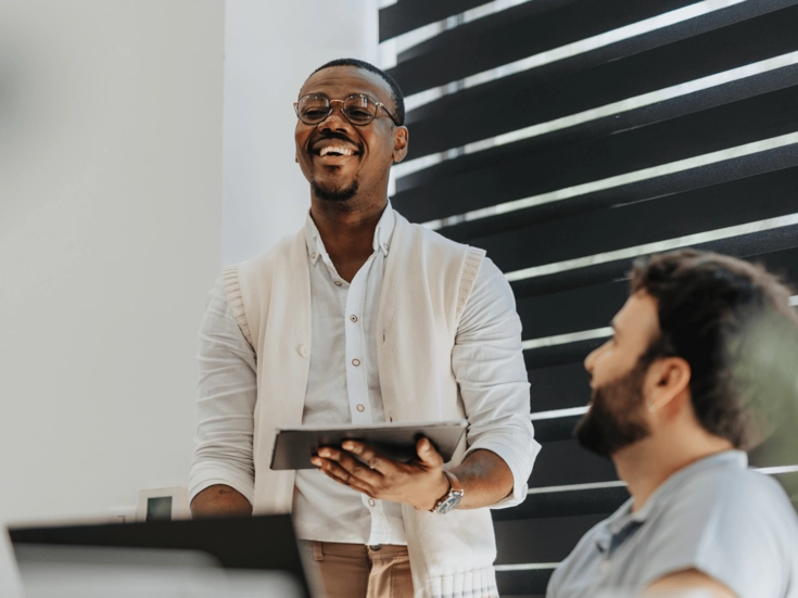 Junger Mann steht lächelnd vor seinem Team und hält ein Tablet während einer Besprechung.