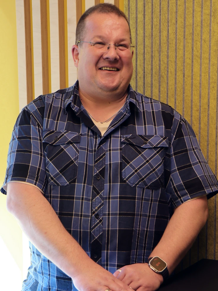 Portrait eines lächelnden conet Mitarbeiters im karierten Kurzarmhemd, entspannt stehend in gelbem Meetingraum 