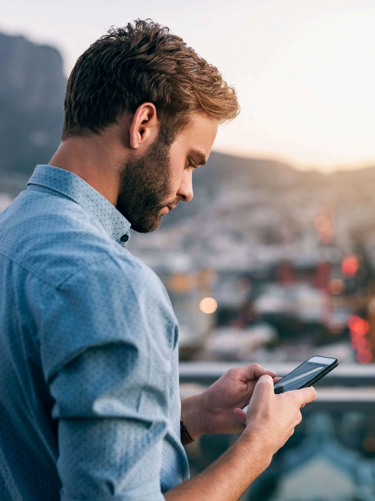 Mann steht auf einer Dachterrasse und nutzt sein Smartphone mit Blick über eine Stadtlandschaft.