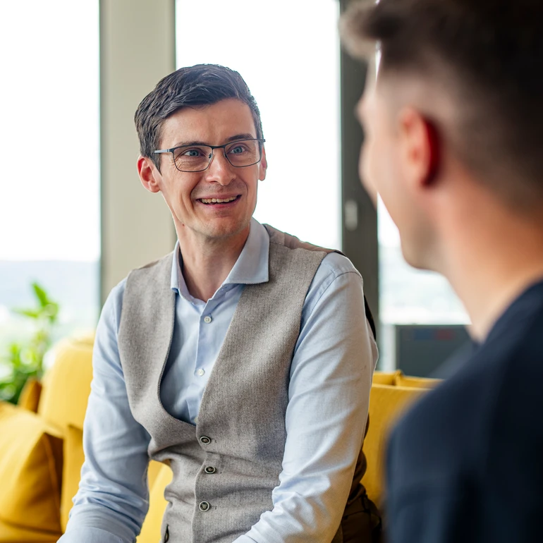 Offenes Gespräch im Büro IT-Consultant in hellblauem Hemd und hellbrauner Weste im Gespräch mit Werkstudent in dunklem T-Shirt