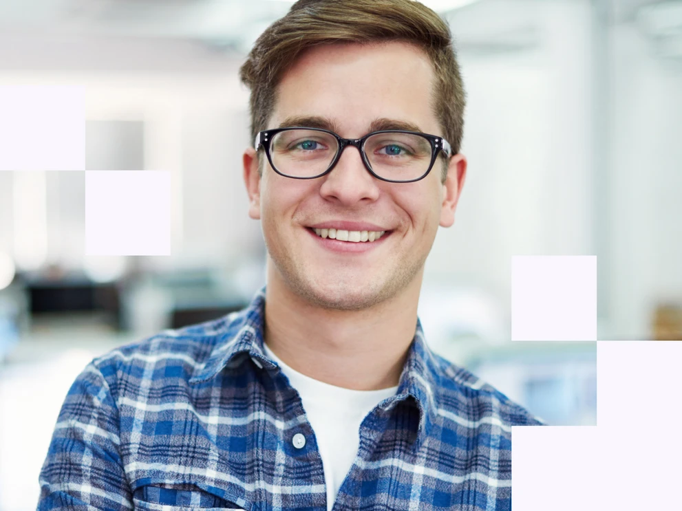 Junger Mann mit Brille im karierten Hemd lächelt in die Kamera, heller Büroraum im Hintergrund